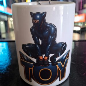 Mug TOY 450 ml avec personnage en combinaison brillante de style puppy et latex, accroupi au-dessus du mot TOY. Design inspiré de l'univers fetish et de la soumission.